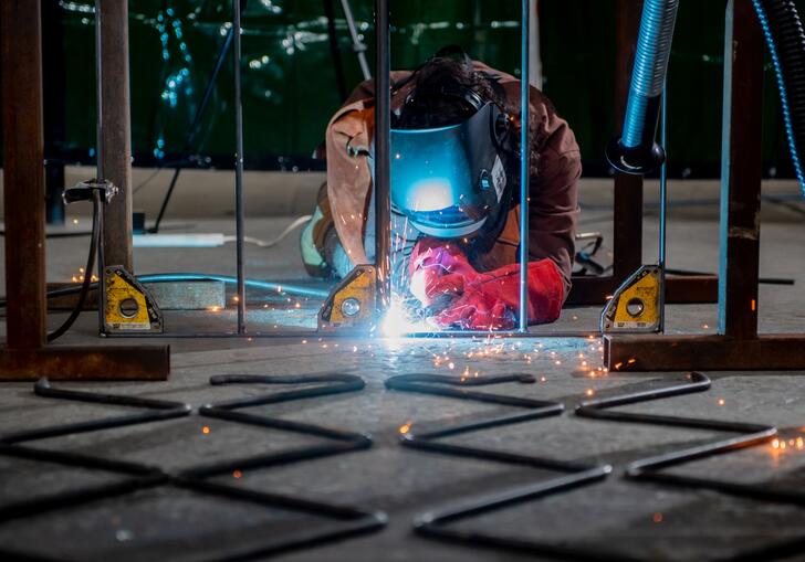 Performance artist Rachel Mars lies on the floor melding a metal gate. She wears protective clothing, including a metal helmet. 