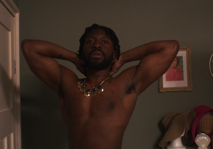 A topless man wearing a large, blingy necklace checks himself out in a mirror in a dark bedroom