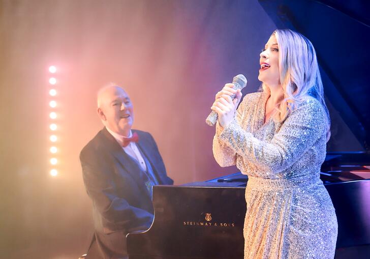 Louise Dearman singing into a microphone, with pianist Jonathan Cohen on piano behind her