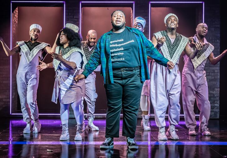 The company of a strange loop pose on stage. A person wearing a green and blue chequered shirt stands front and centre, as six other performers sing and pose behind them.