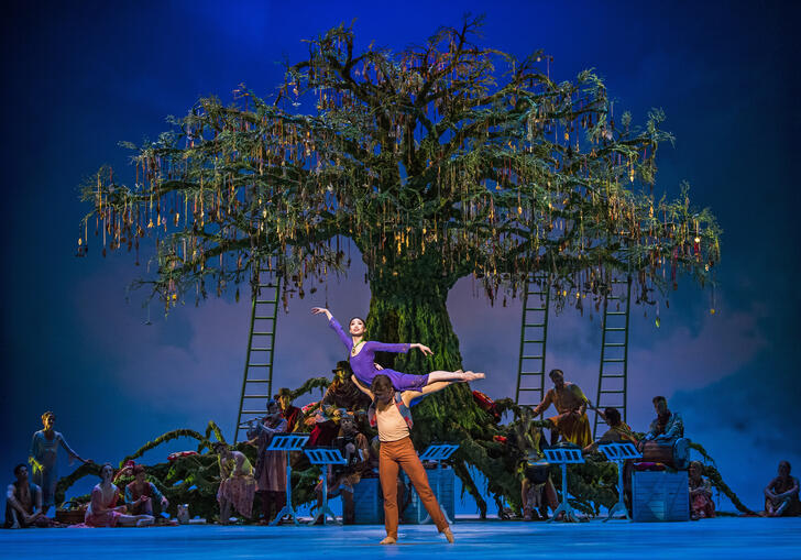 A dancer is lifted into the air by their co-performer on a stage with a large tree