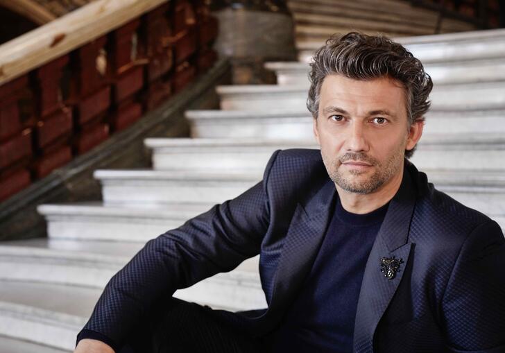 Jonas Kaufmann sits on marble steps in a dark suit 