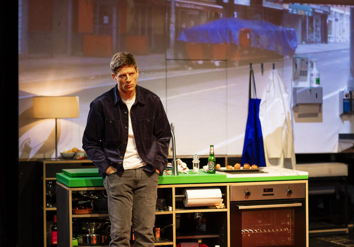 A man looks sad as he leans against a kitchen table on stage