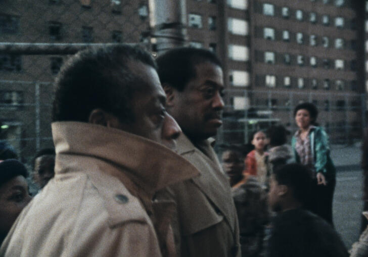 James Baldwin stands on a built up road with his jacket collar around his face