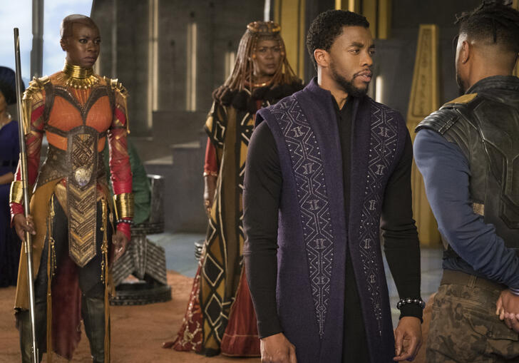 A group of regal people stand in a sci-fi style room, looking serious.