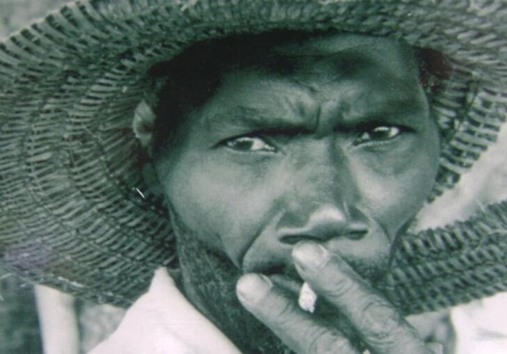 A man wearing a wide brimmed hat smokes a cigarette and stares directly into the camera