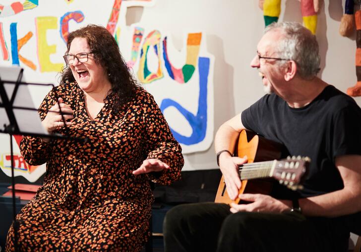 Two individuals, one is playing guitar and another looks on smiling