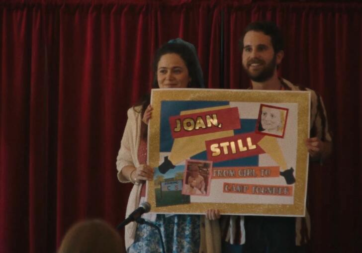 Two young adults hold a homemade sign that reads 'Joan Hill' in front of a red theatre curtain.