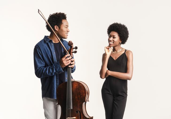 Sheku and Isata Kanneh-Mason smiling at each other. Sheku is holding his cello and bow and they are standing in front of a white background.