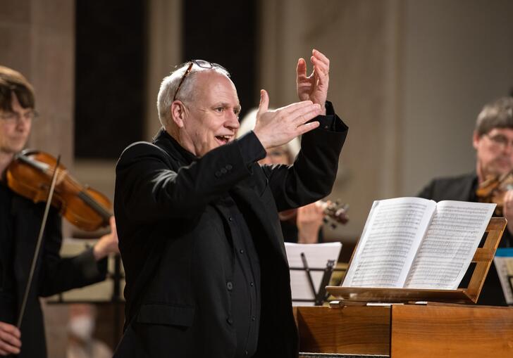 Musical Director John Butt standing by a harpsichord with his arms raised. In the background are two violinists.