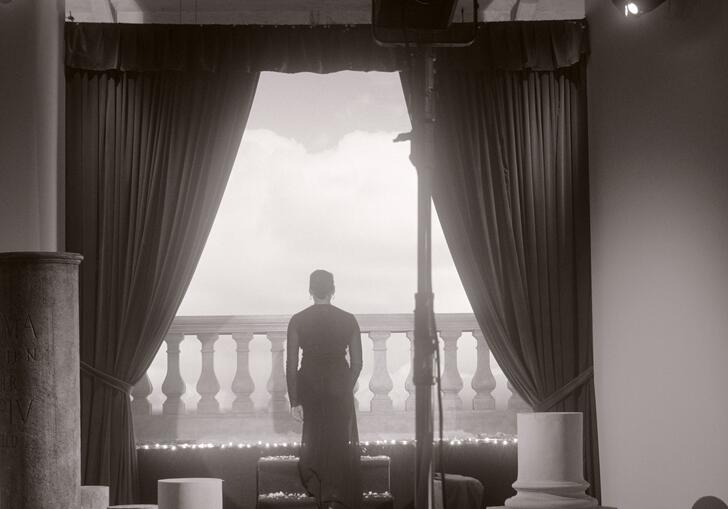 Black and white image of a woman standing with her back to the camera, looking out on to a balcony
