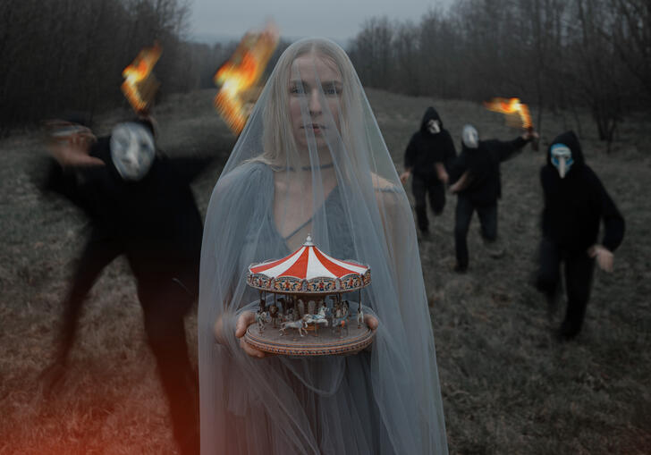 A person wearing a grey veil stands on grass holding a miniature carousel while a group of people dressed in black and wearing haunting facemasks run forwards holding fire.