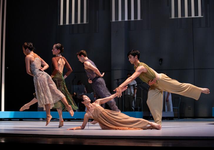 An ensemble of five dancers move and pose elegantly in muted clothing. At the forefront of the stage, two dancers clasp hands. 