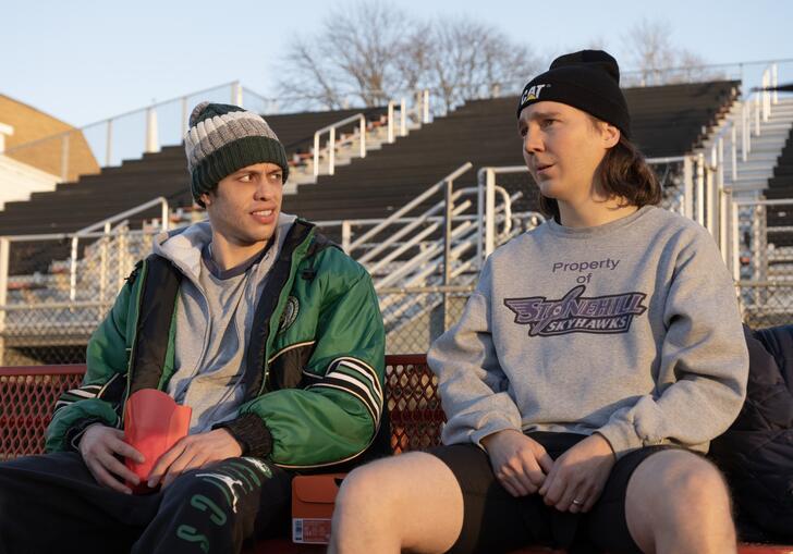 Two young men sit on bleachers looking at each other.
