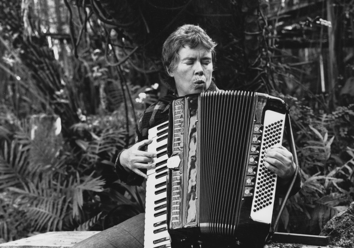 A woman sits on a bench outside and plays an accordion. 