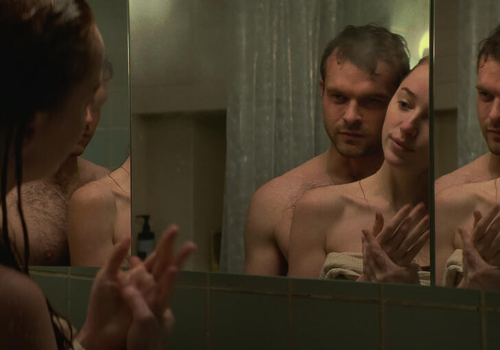 A man and a woman hold each other in a bathroom, seen from a mirror. 