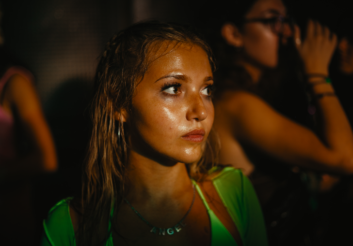 A young woman in a sweaty club looks forward, dazed, whilst bodies dance around her in the dark.