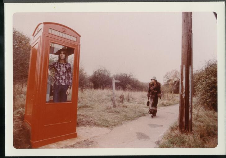 A man stands in a red phone booth on a rural path in an old 1970s photograph. 