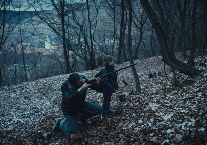 A dad points a gun forward whilst standing next to his young son in a forest at nightfall. 