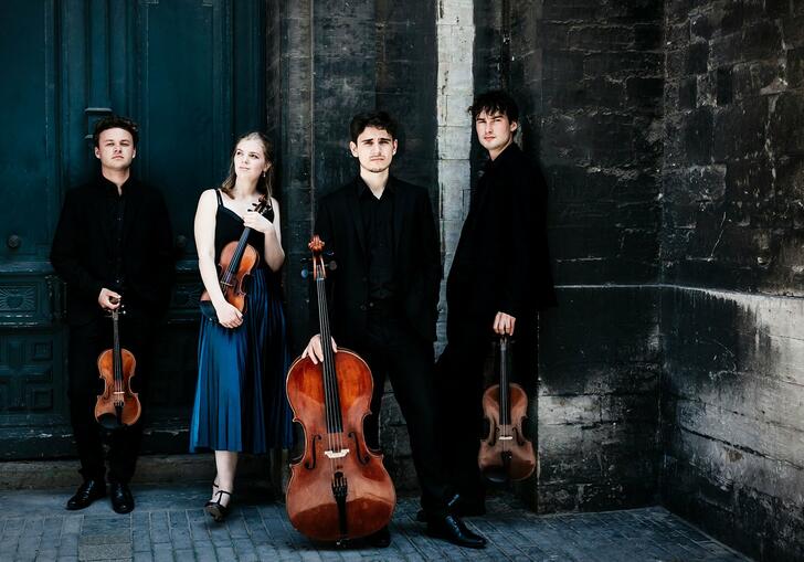 The Sonoro Quartet standing in front of a stone wall and large blue door, holding their instruments