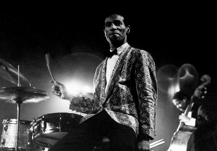 A man in a suit stands by a drum kit in a black and white image