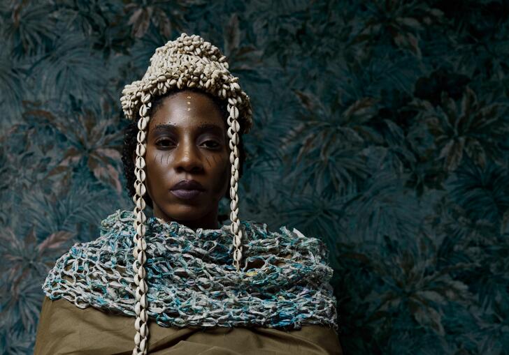 Performer Julene Robinson stands against a blue floral background. She wears cream and blue chains around her head and shoulders.