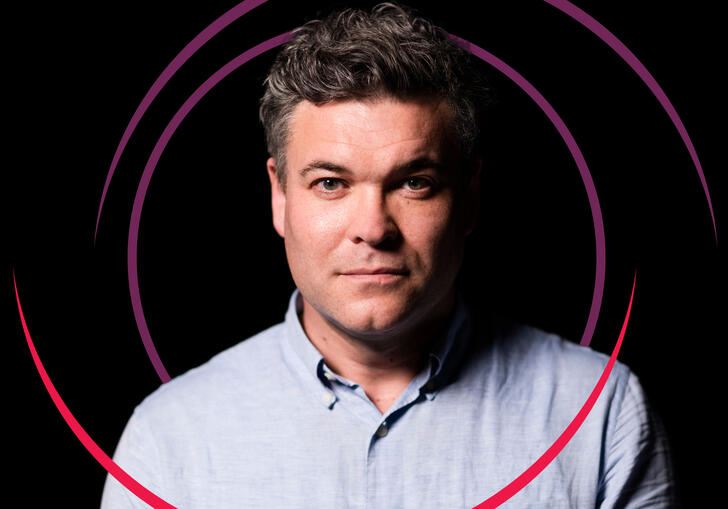 A picture of Ryan Wigglesworth who wears a blue shirt and stands against a black background. Two circles swirl around him in different shades of pink.