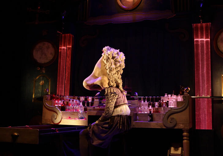 A person with a large nose as a head sits at a table that is filled with perfume bottles.