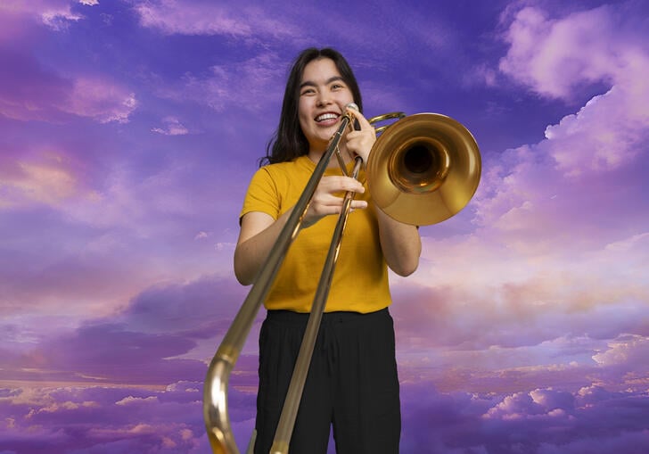 A trombonist from The National Youth Orchestra holding her trombone in front of a skyscape background