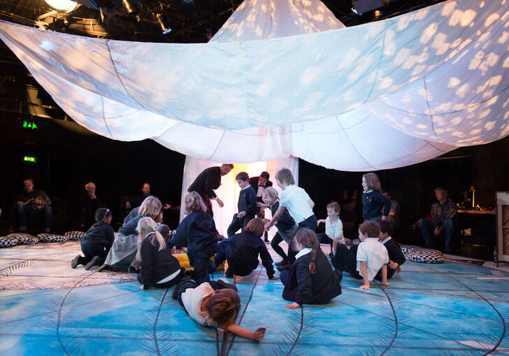 A group of families enjoying a performance of Told by an Idiot's Get Happy. Children gather under a big, white parachute. 