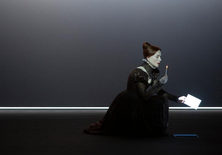 A person dressed as Mary Stuart kneels on the floor in an old-fashioned gown with a lighted match in their hand and a letter in the other.