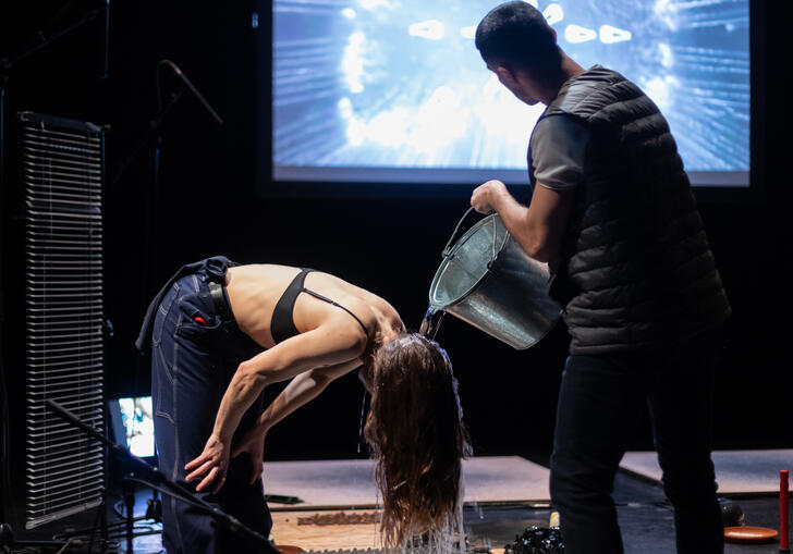 A person pours water over the head of another person as they stand on stage. A psychedelic image is projected in the background.