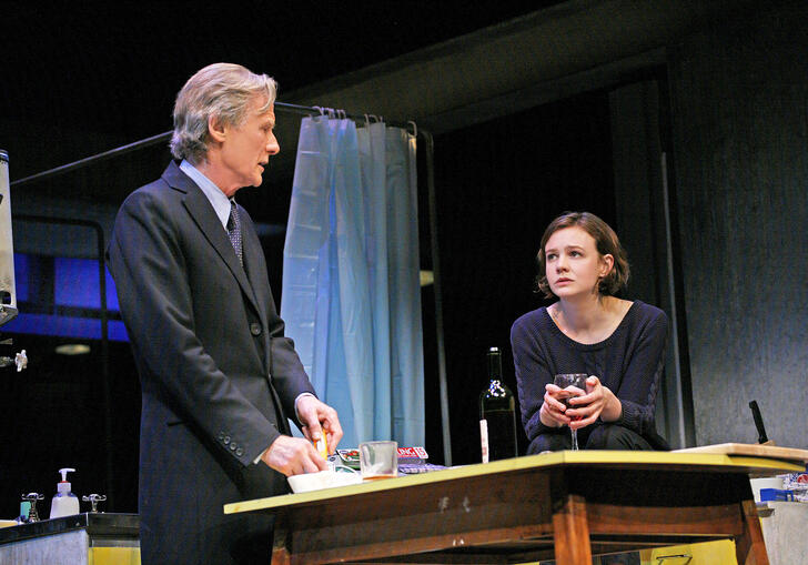 A man and a woman look at each other over a kitchen table, on a theatre stage. 