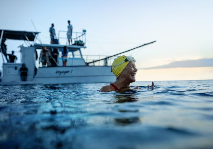 A swimmer with a swimming cap on smiles in the ocean, in front of a white boat.