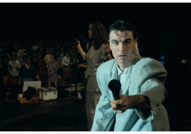 A man holds a microphone out to the camera on a dark stage.