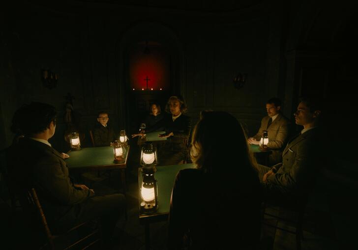 A group of suited people sit in a dark, candlelit room around a table.