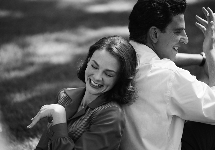 A black and white photograph of a man and a woman resting against each other's backs in a park, laughing together.