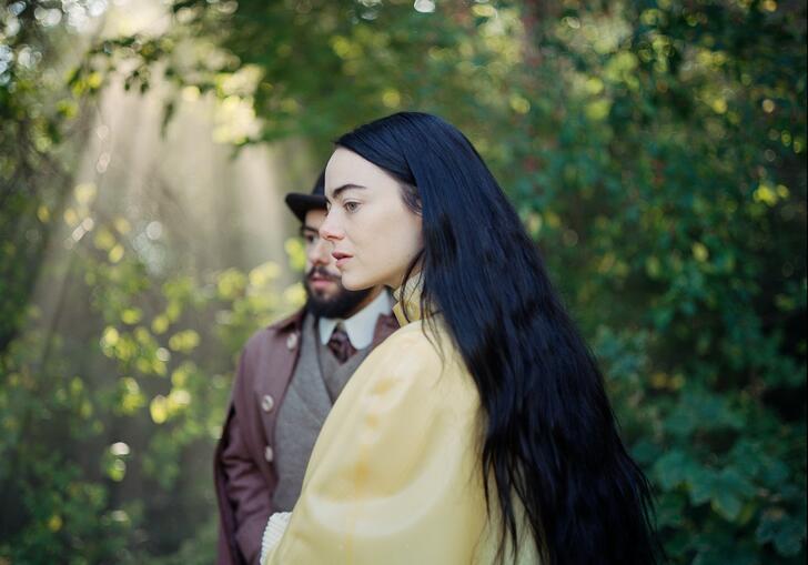 A woman in a yellow dress stands in front of a man in a forest.