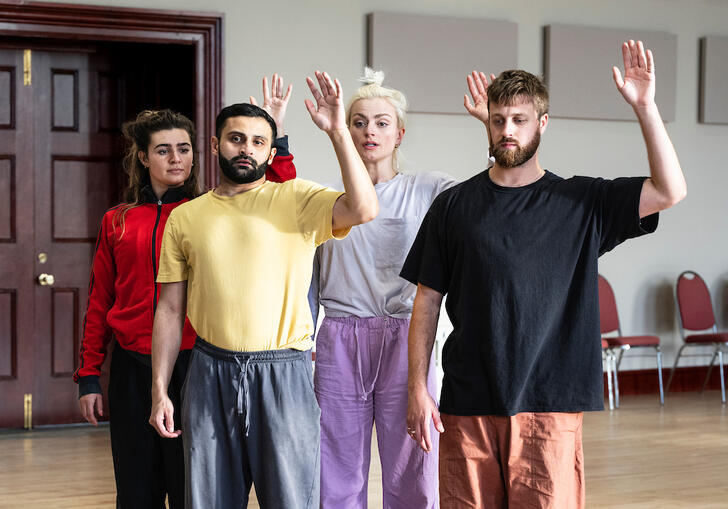 Four people stand in a group with one hand raised in the air as if ready to wave. They have concerned expressions on their faces.