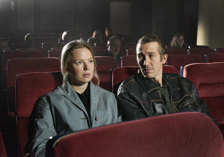 A man looks at a woman who looks straight ahead whilst they sit in a dark cinema.