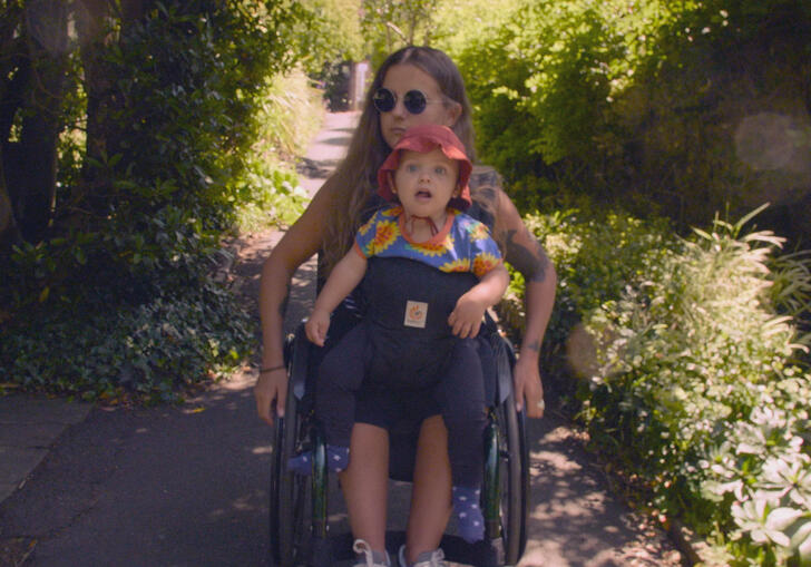 A mother in a wheelchair carries her young child through a flower lined path. 