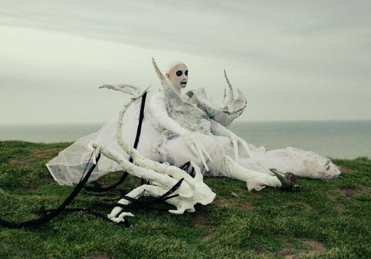 A performance artists lies on the grass of a clifftop, dressed in costume. 