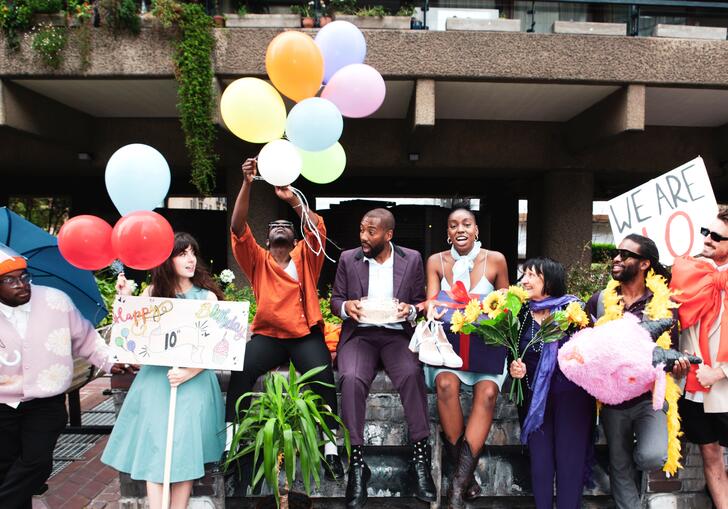 The PappyShow company sit on a wall at the Barbican Centre with signs, balloons, flowers and other objects celebrating the fact they are 10.