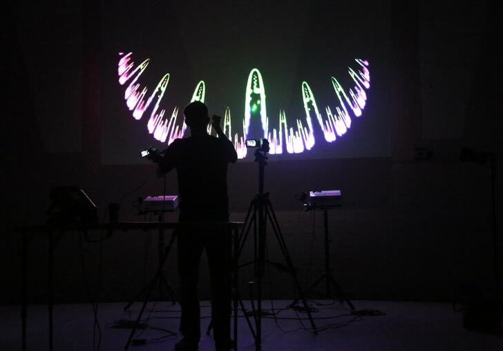 A person stands in a dark room surrounded by technical equipment. A swirling purple and green line is projected before them on a screen.