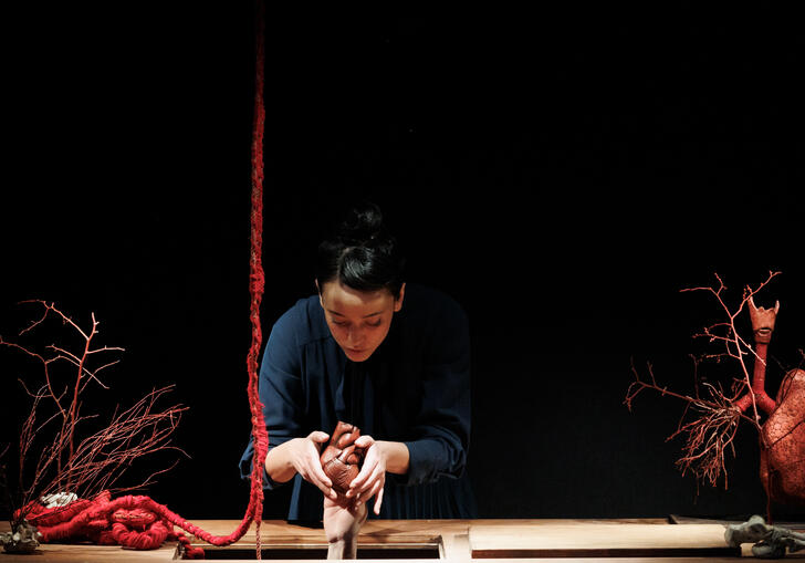 A person stands at a wooden work bench and moulds a small human body out of clay. A large red rope hangs from the ceiling like intestines. 