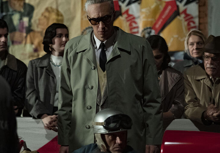 Enzo Ferrari wears glasses on a busy street, in front of a race car.