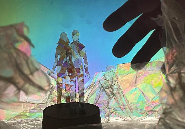 Two figures stand facing each other, they look like they are made out of colourful and transparent material. A large hand reaches towards them.