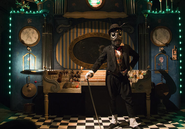 A person dressed as an insect, wearing a suit and holding a cane, stands in front of a large perfume cabinet.