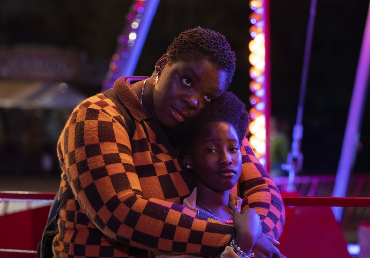 A mother hugs her daughter from behind, standing in front of a bright ferris wheel.