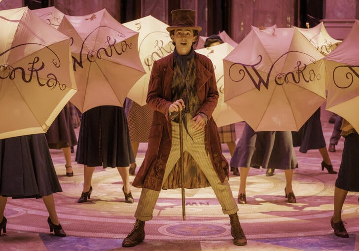 Willy Wonka, wearing a top hat and cane, stands on stage surrounded by umbrellas with 'WONKA' written on them.